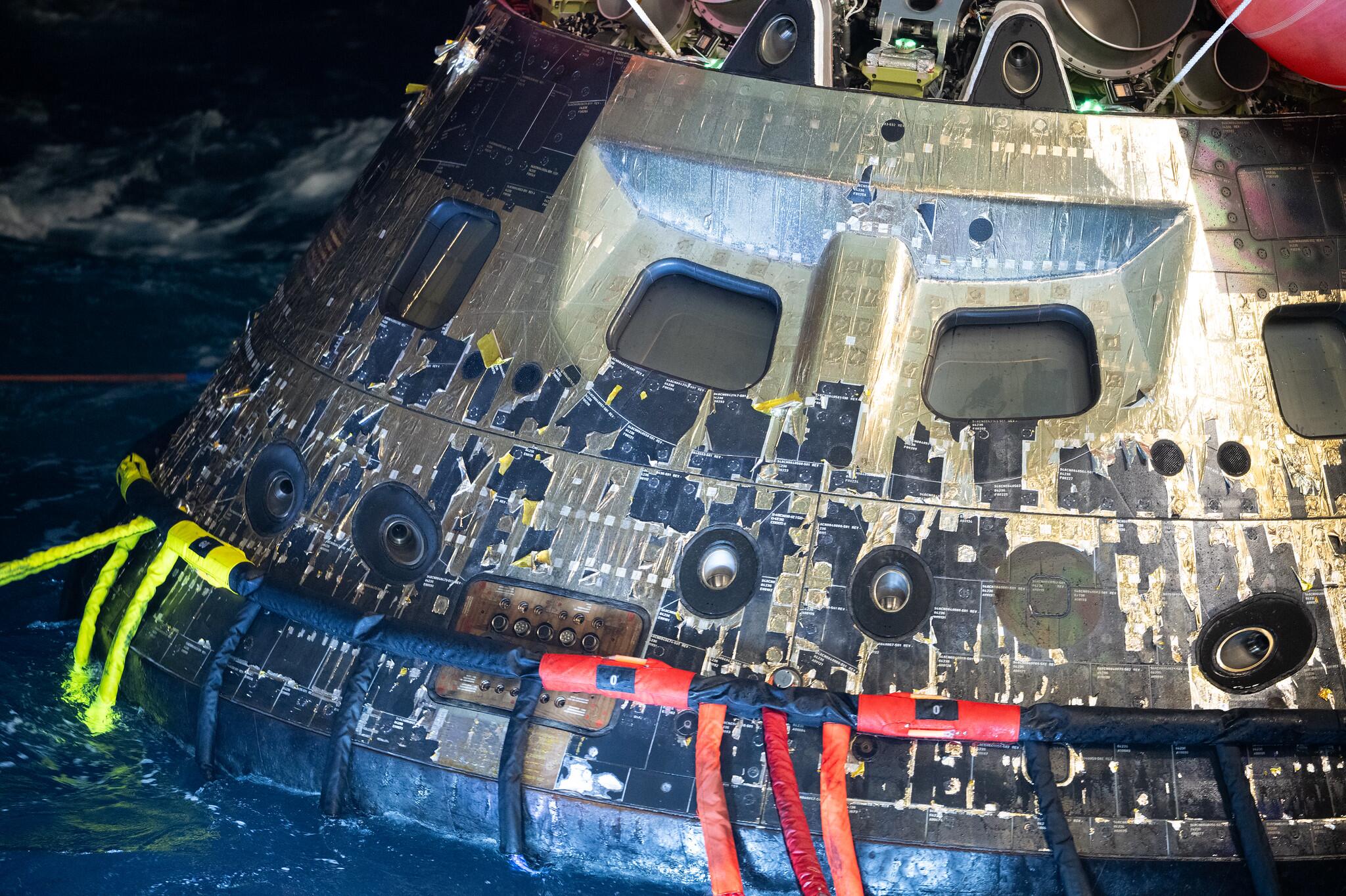 Cápsula espacial flutuando parcialmente na água do mar, com a parte inferior em contato com a superfície e cabos coloridos — amarelos, vermelhos e pretos — conectados à estrutura. A cápsula apresenta revestimento externo em placas metálicas com marcas de desgaste, manchas e áreas escurecidas, além de janelas pequenas e circulares distribuídas pela superfície. Ao fundo, a água aparece em tom escuro, com reflexos e ondulações ao redor da cápsula.