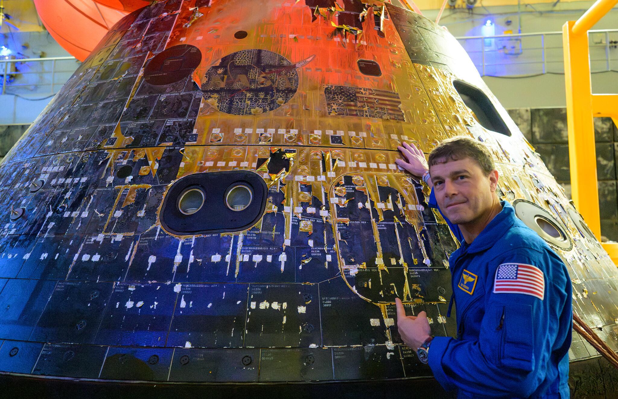 Homem de pele clara, cabelo curto castanho, usando macacão azul com um patch da bandeira dos Estados Unidos no braço, em pé ao lado de uma cápsula espacial. Ele apoia uma das mãos na superfície da cápsula e aponta para uma área do revestimento externo com a outra mão. A cápsula apresenta placas metálicas com diferentes tons, marcas de desgaste e áreas escurecidas, além de pequenas janelas circulares. O ambiente é interno, com iluminação artificial e estruturas ao fundo que sugerem um museu ou instalação técnica.