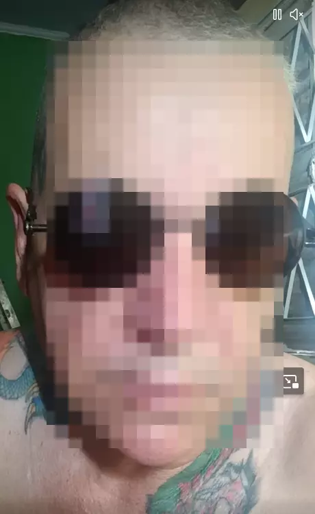 Imagem mostra rosto de homem de pele branca, careca e sem camiseta. O rosto do homem está pixelado para esconder sua identidade, mas é possível identificar que ele usa óculos escuros redondos e tem uma tatuagem de dragão no pescoço.