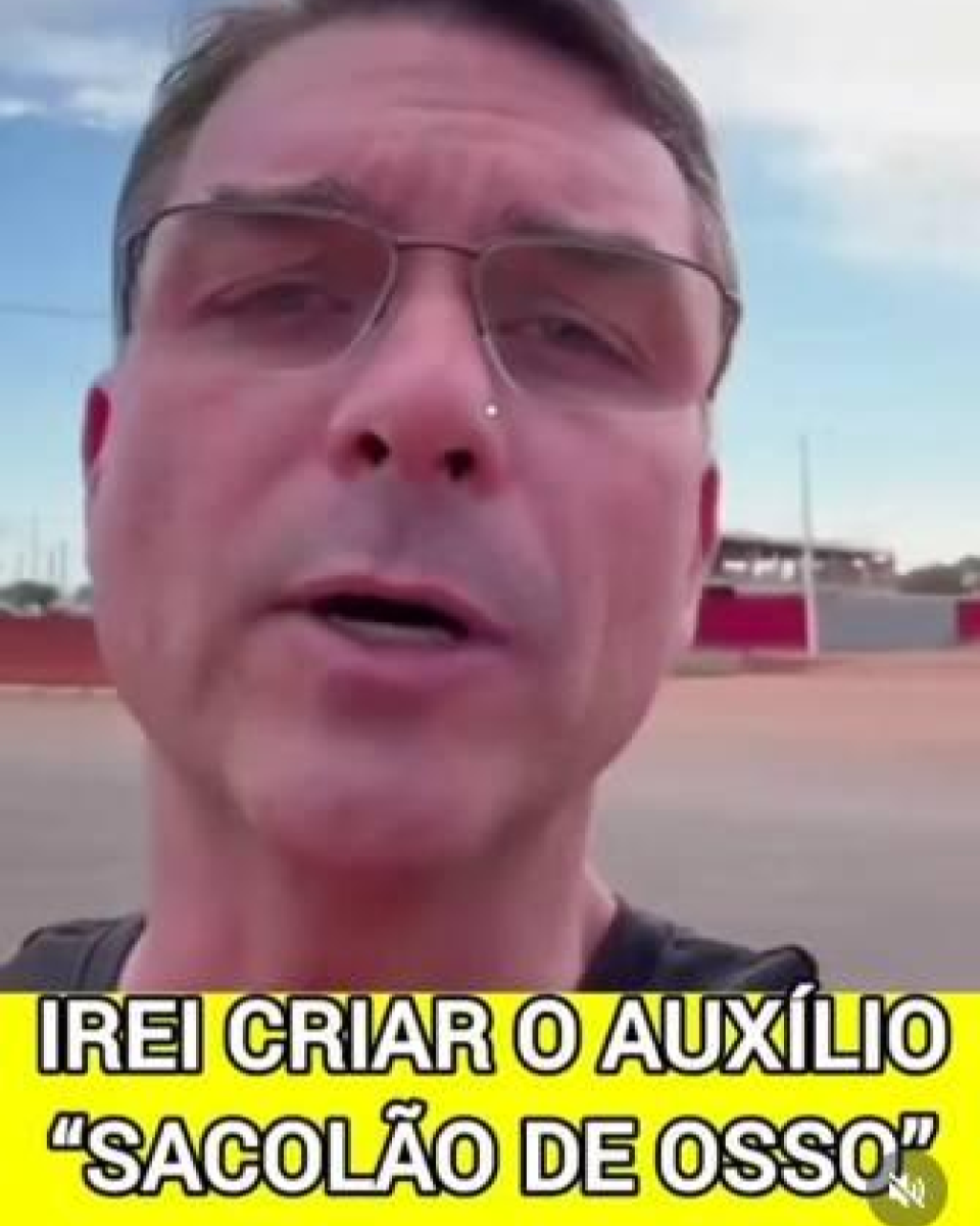 A imagem mostra Flávio Bolsonaro, um homem de pele clara, com cabelo curto castanho e usando óculos de armação fina, em um enquadramento de selfie ao ar livre, com céu parcialmente nublado ao fundo e uma área aberta com construções baixas. Ele veste camiseta escura e olha diretamente para a câmera com expressão séria. Na parte inferior da imagem, há uma faixa amarela com texto em destaque que diz: ‘IREI CRIAR O AUXÍLIO “SACOLÃO DE OSSO”’. 