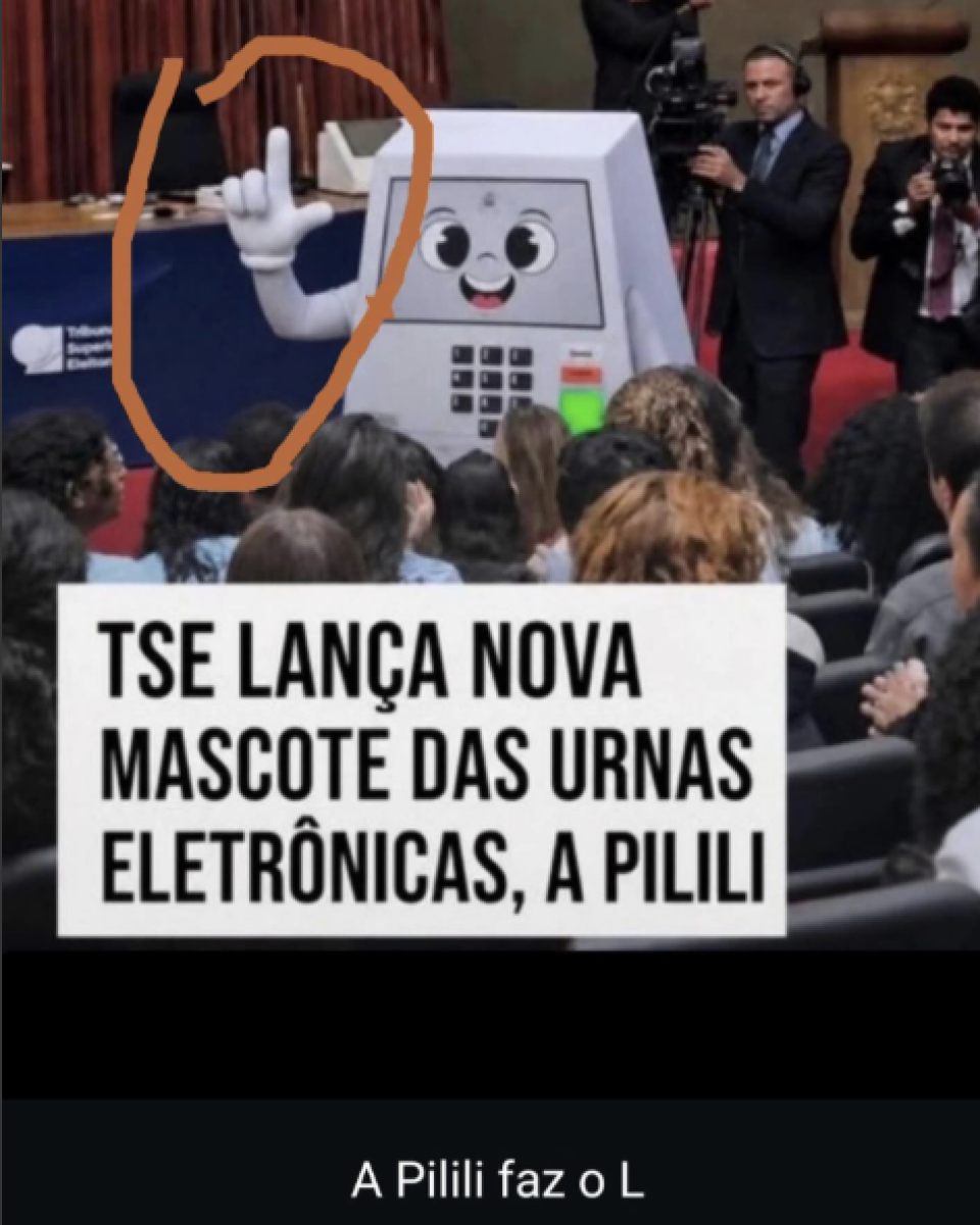 A imagem mostra um auditório cheio de pessoas sentadas, vistas de costas. Na frente da sala está Pilili, uma personagem fantasiada de urna eletrônica com rosto desenhado na tela, olhos grandes e sorriso. Ela tem braços brancos e mostra uma das mãos levantada, fazendo um gesto com o polegar e o indicador estendidos. Em volta desse braço há um círculo desenhado em laranja. Ao fundo, aparecem dois homens de terno segurando câmeras. Na parte inferior da imagem, há uma caixa branca com texto em letras pretas dizendo: ‘TSE LANÇA NOVA MASCOTE DAS URNAS ELETRÔNICAS, A PILILI’. Abaixo da imagem principal, sobre um fundo preto, aparece a frase ‘A Pilili faz o L’ em letras brancas.