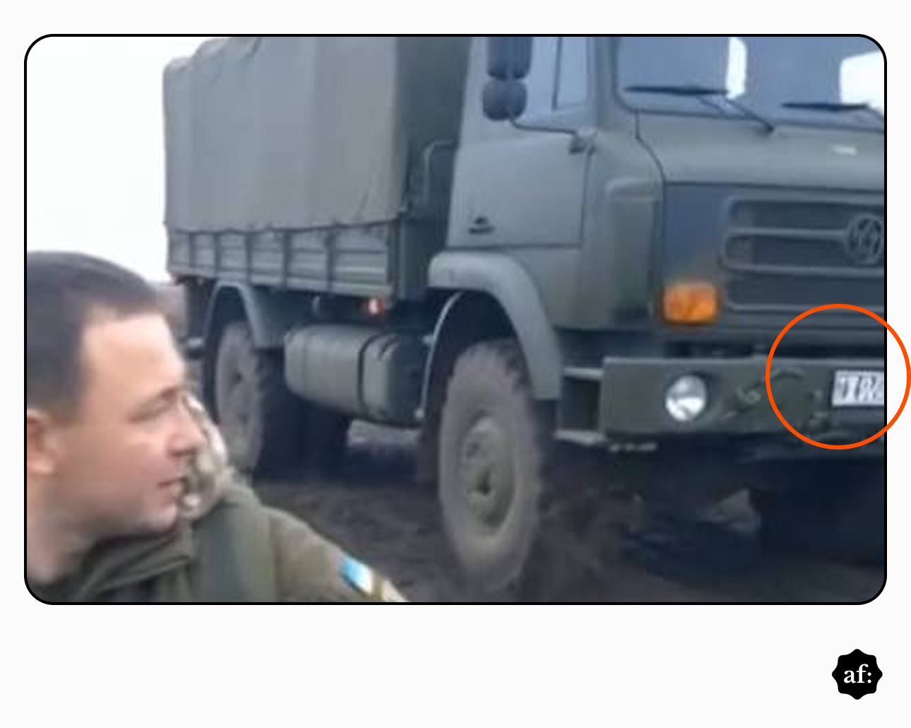 Captura de tela mostra rosto de homem com trajes militares do lado esquerdo e caminhão militar verde estacionado em área de terra ao fundo. Um círculo laranja destaca a placa dianteira do veículo, que exibe caracteres borrados e ilegíveis, com um formato diferente do padrão oficial.