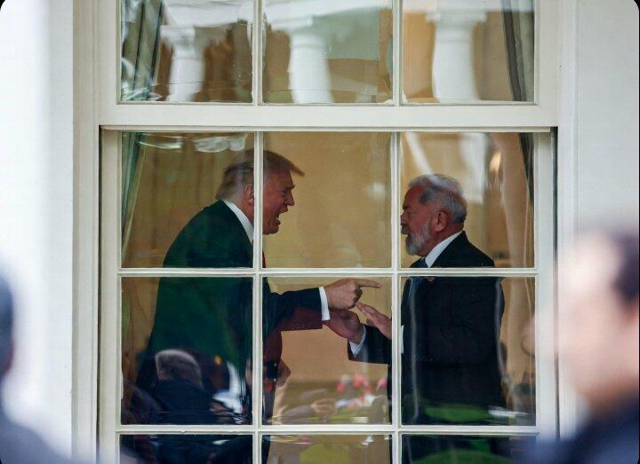 Imagem mostra dois homens de pele clara vistos através de uma janela com divisórias brancas, em um ambiente interno iluminado. À esquerda, Donald Trump, homem com cabelo claro penteado para trás, usando terno escuro, aparece inclinado para frente enquanto aponta o dedo em direção ao outro homem. À direita, Lula, um homem com cabelo curto branco e barba grisalha, também usando terno escuro, gesticula com as mãos enquanto olha para o interlocutor. O cenário ao fundo possui cortinas claras e móveis pouco visíveis, enquanto áreas desfocadas aparecem nas laterais externas da janela.