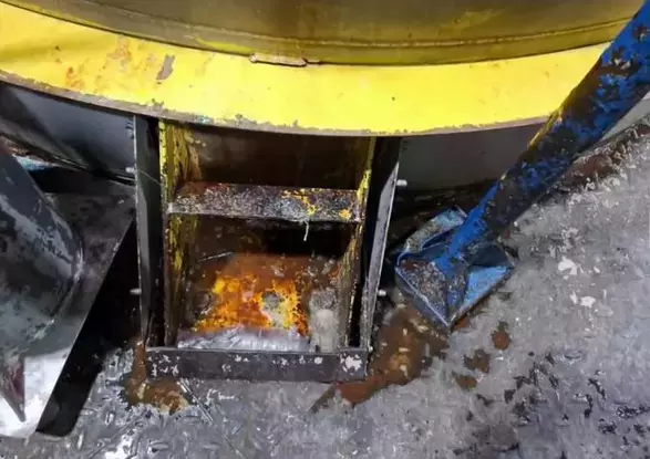 Close em uma estrutura metálica enferrujada. Na parte superior da imagem, há uma superfície curva amarela com sinais de desgaste e ferrugem. No centro, aparece um compartimento metálico oxidado com resíduos acumulados. À direita, uma peça azul metálica também apresenta desgaste. O chão está com aspecto corroído.