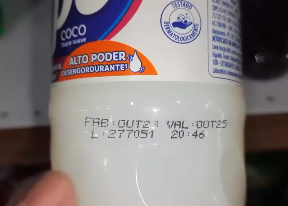 Close em embalagem branca de detergente de coco da Ypê. O rótulo exibe frases como ‘ALTO PODER DESENGORDURANTE’ e selo de ‘TESTADO DERMATOLOGICAMENTE’. Na parte inferior da embalagem aparecem informações impressas em tinta preta com dados de fabricação e validade, incluindo ‘FAB: OUT23’ e ‘VAL: OUT25’, além de um código de lote e horário de produção. Um dedo aparece apontando para a área onde as informações estão impressas.