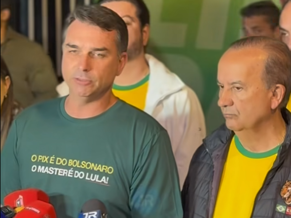 Duas pessoas aparecem em primeiro plano durante uma coletiva de imprensa. À esquerda, Flávio Bolsonaro — homem de pele clara e cabelo curto castanho, usa camiseta verde com a frase ‘O PIX É DO BOLSONARO O MASTER É DO LULA!’ escrita em amarelo e branco. À direita, Jorginho Mello — homem de pele clara e cabelo curto grisalho — usa camiseta amarela com gola verde e jaqueta escura. Microfones aparecem na parte inferior da imagem. Ao fundo, há outras pessoas desfocadas, algumas vestindo roupas nas cores verde e amarelo.