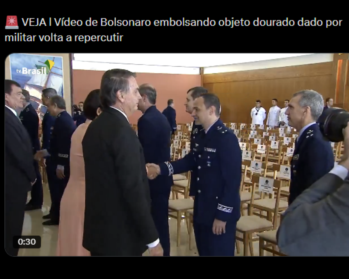  Publicação em rede social com fundo preto. No topo, há um emoji de sirene vermelha seguido do texto: 'VEJA | Vídeo de Bolsonaro embolsando objeto dourado dado por militar volta a repercutir'. Abaixo, aparece a miniatura de um vídeo com duração de '0:30', indicada no canto inferior esquerdo. A cena mostra um salão amplo com várias cadeiras de madeira organizadas em fileiras. No centro da imagem, Jair Bolsonaro, vestindo terno escuro, aperta a mão de um militar usando uniforme azul da aeronáutica. Outro militar com uniforme semelhante está à direita, observando a cena. Há outras pessoas ao fundo e uma pessoa segurando uma câmera aparece parcialmente no lado direito da imagem. No canto superior esquerdo da miniatura do vídeo há um logotipo escrito 'TV Brasil'.