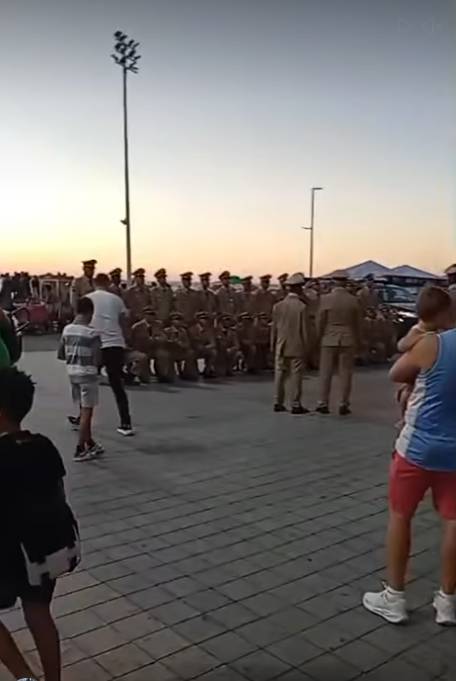 Foto tirada ao ar livre ao entardecer mostrando um grande grupo de militares uniformizados reunidos em formação para uma foto coletiva. Os agentes usam uniformes cáqui e bonés, alguns em pé e outros agachados na frente do grupo. No centro da cena, um homem de camiseta branca e calça escura parece fotografar ou organizar os participantes. Ao redor, pessoas observam a movimentação, incluindo uma criança à esquerda e uma mulher de shorts rosa à direita segurando um celular. O chão é pavimentado com blocos cinza e o céu aparece claro no horizonte. 