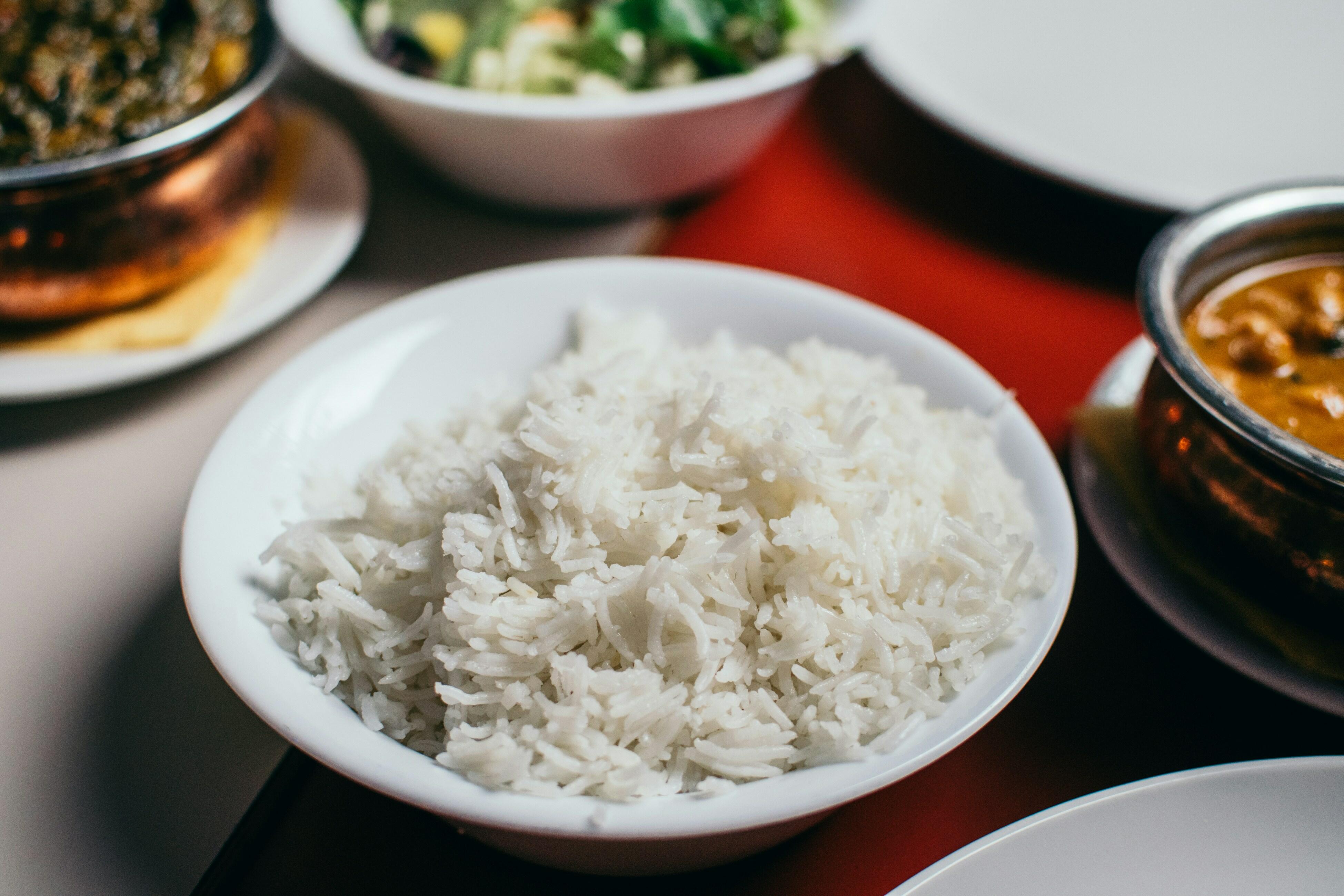 Em primeiro plano, há uma tigela branca cheia de arroz branco cozido, com os grãos soltos e levemente brilhantes. A tigela está sobre uma mesa de superfície clara. Ao redor dela, aparecem parcialmente outros recipientes com alimentos: à esquerda, um bowl metálico com um prato escuro; ao fundo, uma tigela branca com vegetais verdes e pedaços claros; à direita, um recipiente metálico com um ensopado de cor amarelada; e, no canto inferior direito, a borda de um prato branco. Sobre a mesa, há um tecido vermelho que passa por trás da tigela de arroz e se estende até o lado direito da imagem. O foco da fotografia está concentrado na tigela de arroz, enquanto os demais elementos aparecem desfocados.