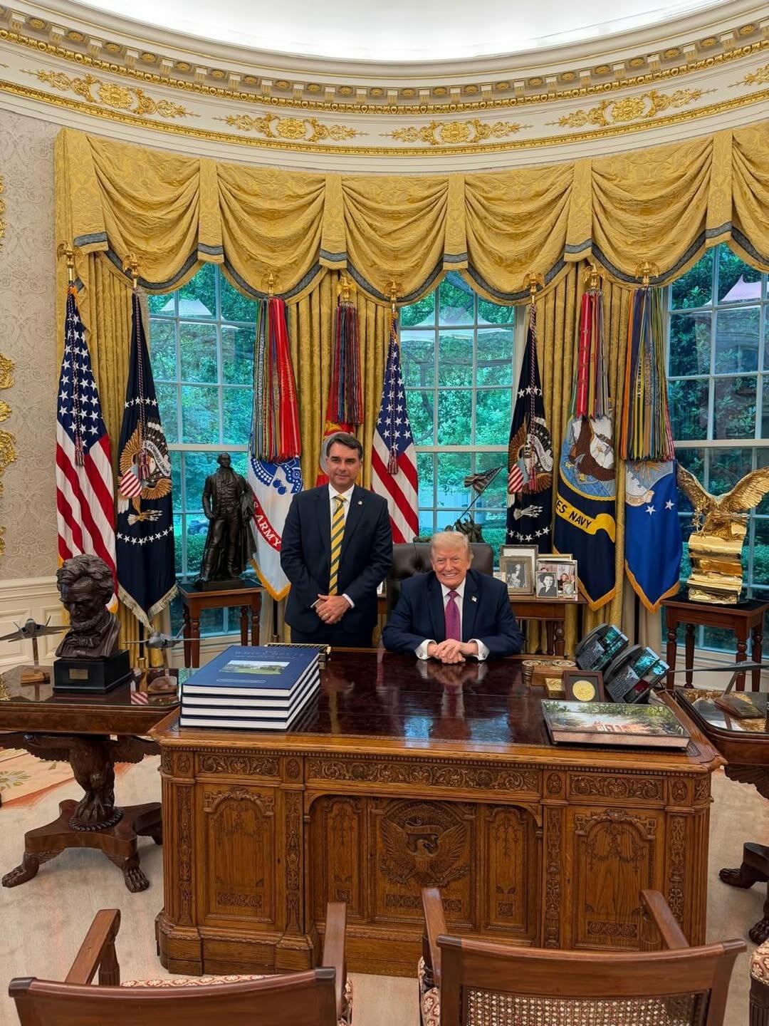 Imagem mostra o presidente Donald Trump — homem de pele clara, cabelo loiro penteado para trás, usando terno escuro e gravata vermelha — sentado atrás da mesa presidencial no Salão Oval da Casa Branca. Ao lado dele está Flávio Bolsonaro — homem de pele clara, cabelo curto escuro, usando terno azul-marinho, camisa branca e gravata amarela — em pé com as mãos cruzadas à frente do corpo. Sobre a mesa há pilhas de livros, telefones, molduras com fotografias e outros objetos decorativos. Ao fundo aparecem bandeiras dos Estados Unidos e de forças militares, cortinas douradas, janelas com vista para árvores e esculturas posicionadas nas laterais da sala. 