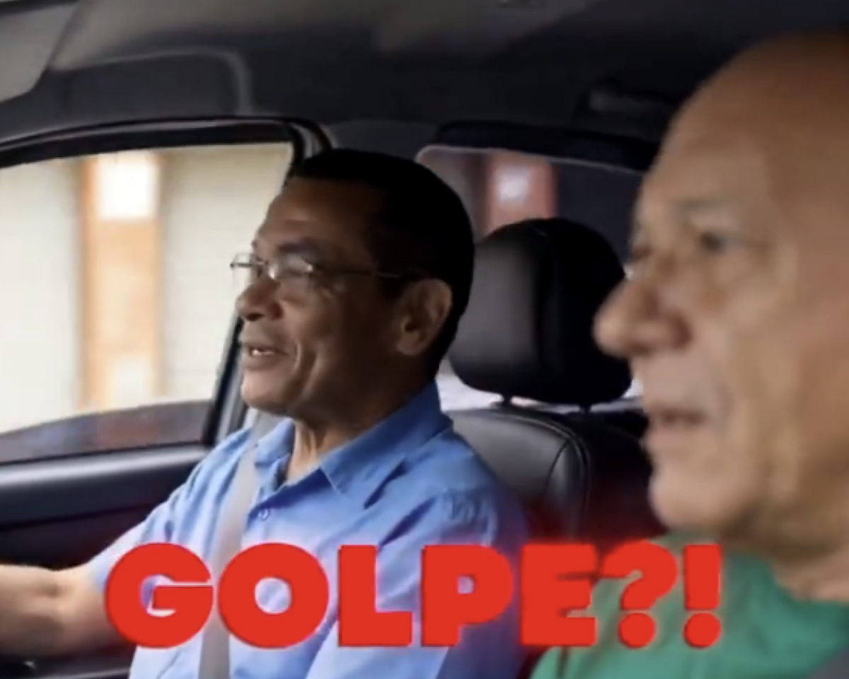 A imagem mostra o interior de um carro visto do banco da frente. Do lado esquerdo, um homem negro que usa óculos e camisa azul clara está sentado no banco do motorista e olha para frente com um leve sorriso. Do lado direito, em primeiro plano e desfocado, aparece o empresário Luciano Hang, careca e de pele clara, usando camiseta verde, sentado no banco do passageiro. Sobre a parte inferior da imagem há a palavra ‘GOLPE?!’ escrita em letras grandes vermelhas. O interior do veículo é escuro, com bancos pretos e partes da janela visíveis ao fundo.