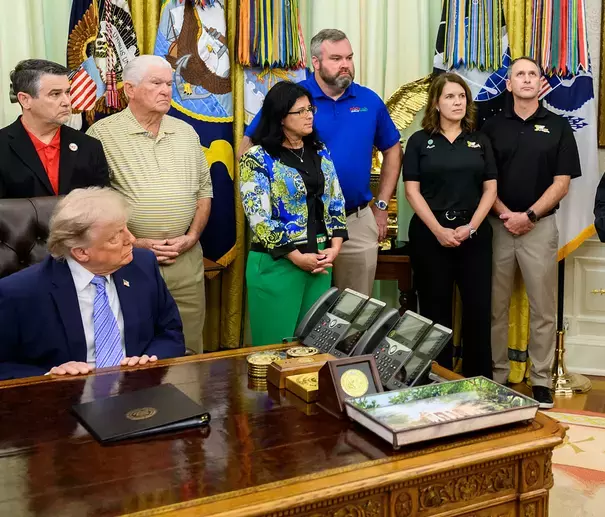 Imagem mostra Donald Trump — homem de pele clara, cabelo loiro penteado para trás, usando terno azul e gravata listrada em azul claro — sentado atrás da mesa presidencial no Salão Oval da Casa Branca, olhando para um grupo de pessoas em pé ao lado da mesa. Entre elas estão homens e mulheres de diferentes idades e tons de pele, usando roupas sociais e camisas polo em cores como preto, azul, vermelho, verde e amarelo. Sobre a mesa aparecem telefones, moedas, porta-retratos, uma pasta preta fechada e uma bandeja com imagem de paisagem. Ao fundo, há bandeiras militares dos Estados Unidos, cortinas douradas e medalhas penduradas.