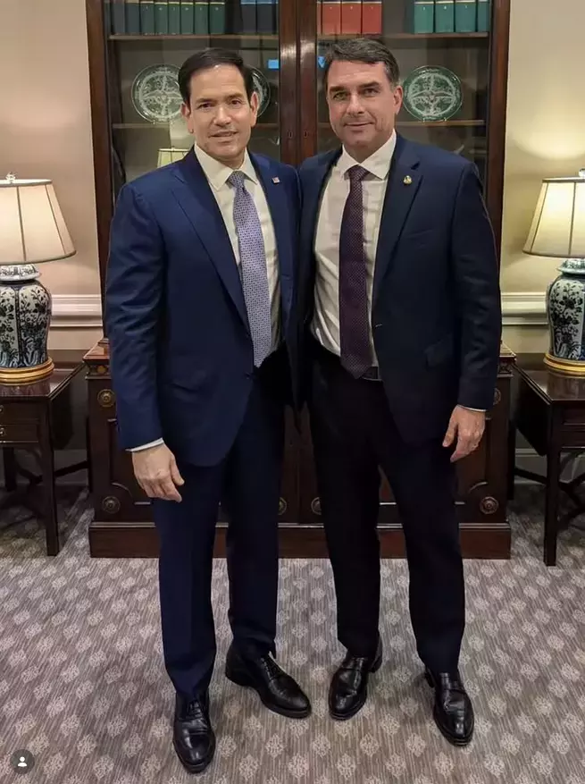 Imagem mostra dois homens de pele clara usando ternos escuros posando lado a lado em um ambiente interno com móveis de madeira escura. À esquerda, está Marco Rubio — homem de cabelo curto escuro, usando terno azul, camisa branca e gravata azul clara com estampa geométrica. À direita está Flávio Bolsonaro — homem de cabelo curto escuro, usando terno azul-marinho, camisa branca e gravata vinho com pequenos pontos claros. Os dois estão de pé sobre um carpete cinza, com os braços próximos ao corpo. Ao fundo, há um armário com portas de vidro contendo pratos decorativos verdes, além de duas luminárias com base azul e branca posicionadas sobre mesas laterais.