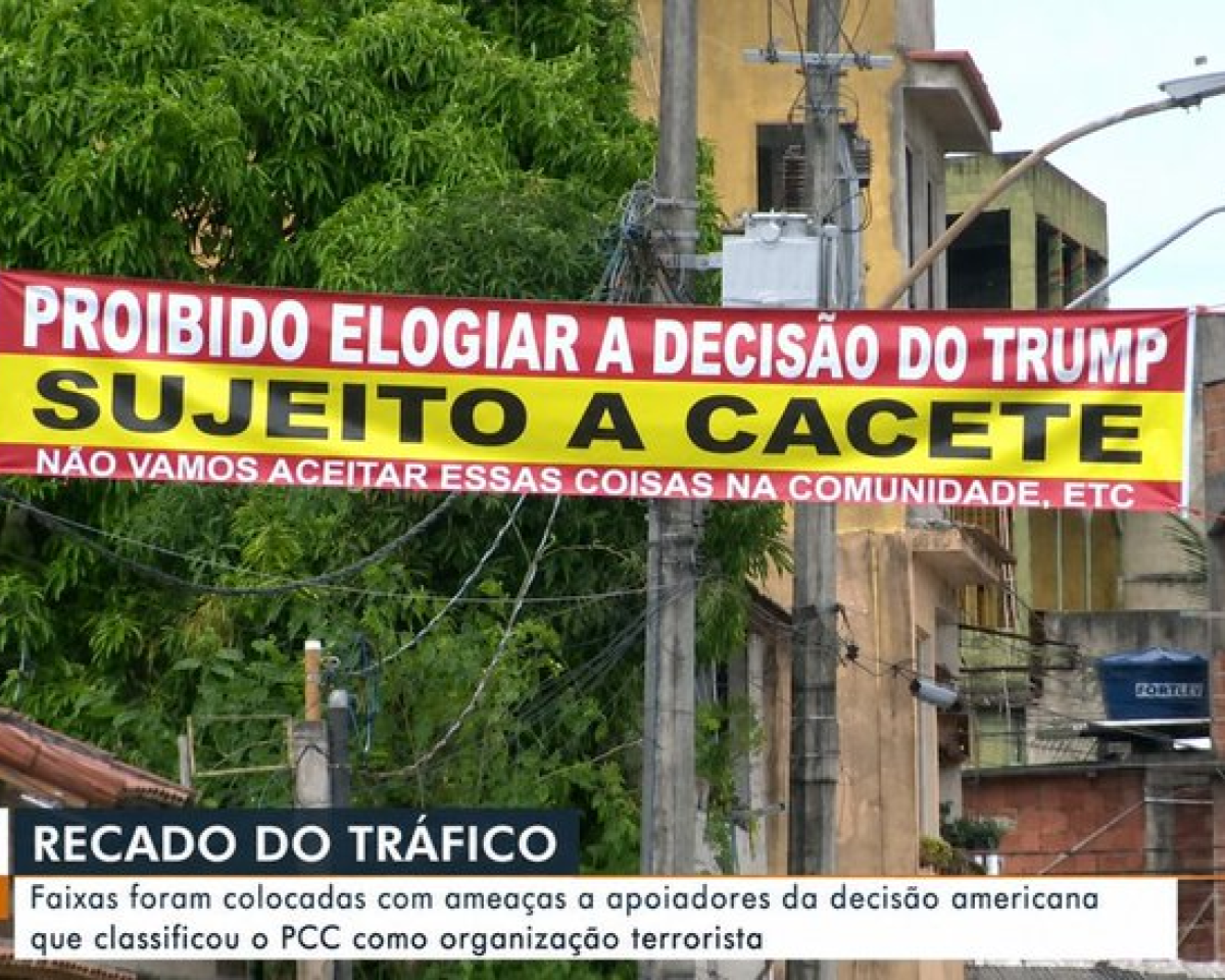 A imagem mostra uma rua com postes, fios elétricos, árvores e construções ao fundo. Há uma faixa horizontal nas cores vermelha e amarela estendida entre dois postes. Na parte superior da faixa, sobre fundo vermelho, está escrito em letras brancas: ‘PROIBIDO ELOGIAR A DECISÃO DO TRUMP’. No centro, sobre fundo amarelo, aparece a frase ‘SUJEITO A CACETE’ em letras pretas grandes. Na parte inferior da faixa, sobre fundo vermelho, está escrito em letras brancas: ‘NÃO VAMOS ACEITAR ESSAS COISAS NA COMUNIDADE, ETC’. Na parte inferior da imagem há a tarja de um telejornal em que aparece o título ‘RECADO DO TRÁFICO’ em letras brancas. Logo abaixo, em uma faixa branca, está escrito: ‘Faixas foram colocadas com ameaças a apoiadores da decisão americana que classificou o PCC como organização terrorista’.