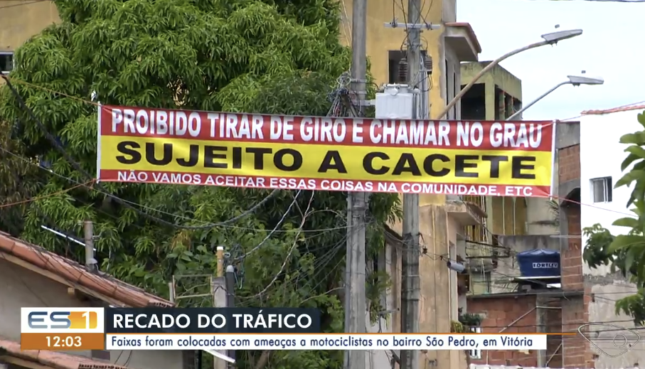 A imagem mostra uma rua com árvores, postes, fios elétricos e edifícios ao fundo. Entre dois postes está estendida uma faixa horizontal nas cores vermelha e amarela. Na parte superior da faixa, sobre fundo vermelho, está escrito em letras brancas: ‘PROIBIDO TIRAR DE GIRO E CHAMAR NO GRAU’. No centro, sobre fundo amarelo, aparece a frase ‘SUJEITO A CACETE’ em letras pretas grandes. Na parte inferior da faixa, sobre fundo vermelho, lê-se: ‘NÃO VAMOS ACEITAR ESSAS COISAS NA COMUNIDADE, ETC’. Na parte inferior da imagem há elementos de uma reportagem de televisão. À esquerda aparece o logotipo ‘ES1’ e o horário ‘12:03’. Ao lado, em uma faixa azul escura, está escrito ‘RECADO DO TRÁFICO’. Logo abaixo, em uma faixa branca, aparece o texto: ‘Faixas foram colocadas com ameaças a motociclistas no bairro São Pedro, em Vitória’. No canto inferior direito há o logotipo de uma emissora de televisão. Ao fundo são visíveis fachadas de casas e prédios, postes de iluminação pública, cabos aéreos e a copa de uma árvore.