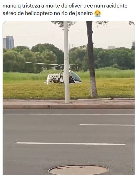 Imagem mostra uma captura de tela de uma publicação em rede social. No topo há o texto: ‘mano q tristeza a morte do oliver tree num acidente aéreo de helicoptero no rio de janeiro’, seguido de um emoji de choro. Abaixo aparece um helicóptero branco e preto parado sobre uma área gramada, próximo a árvores e vegetação. Em primeiro plano há uma via asfaltada com faixas de trânsito, um bueiro e um poste branco que parcialmente encobre a aeronave. Ao fundo são visíveis árvores e edifícios ao longe. Não há pessoas visíveis na imagem. 
