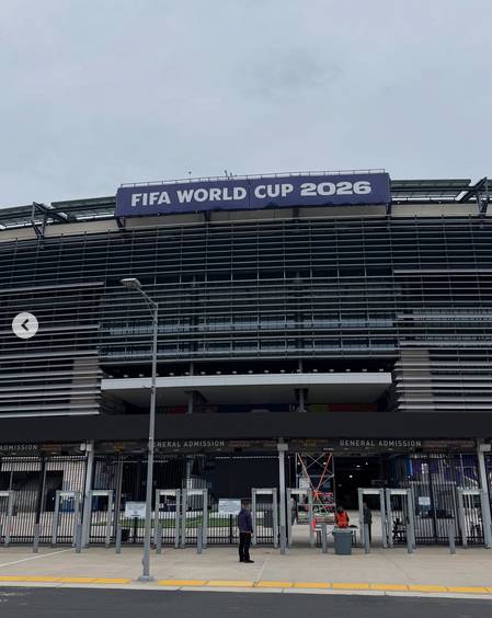 Uma fotografia exibe a vista externa da fachada de um estádio, composta por linhas horizontais sobrepostas, sob um céu nublado. Na parte superior da estrutura, há um painel roxo com os dizeres 'FIFA WORLD CUP 2026' em letras brancas. No nível do solo, encontram-se portões de acesso com detectores de metal e placas pretas onde se lê 'GENERAL ADMISSION' e outras inscrições menores, como 'METAL DETECTORS IN USE'. Há três pessoas distantes próximas às entradas: uma pessoa no centro vestindo roupas escuras, uma segunda pessoa à direita usando roupas escuras e colete refletivo laranja ao lado de uma lixeira cinza, e uma terceira pessoa mais ao fundo, também com vestimentas escuras. Devido à distância e à resolução, não é possível identificar características físicas como cor da pele ou do cabelo dos indivíduos. Um poste de iluminação alto ergue-se à esquerda e, sobreposto à imagem nesse mesmo lado, há um ícone circular cinza com uma seta preta apontando para a esquerda. A calçada apresenta blocos retangulares amarelos em sua borda inferior.