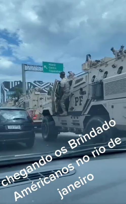 Uma fotografia tirada do interior de um veículo mostra uma cena de trânsito sob um céu parcialmente nublado, onde uma fileira de veículos blindados de cor bege se alinha à direita de um carro de passeio escuro. No blindado mais próximo, que possui a inscrição 'PF' em letras pretas na lateral, há um homem de pele escura, vestindo calça camuflada clara, camiseta clara e boné escuro, posicionado de pé no estribo e segurando a porta aberta do veículo. Acima da via, uma placa de trânsito verde exibe setas e os nomes 'Niterói', 'Centro' e 'Copacabana'. Sobreposta à metade inferior da imagem, há uma frase escrita em letras brancas inclinadas que diz: 'chegando os Brindado Américanos no rio de janeiro'. 