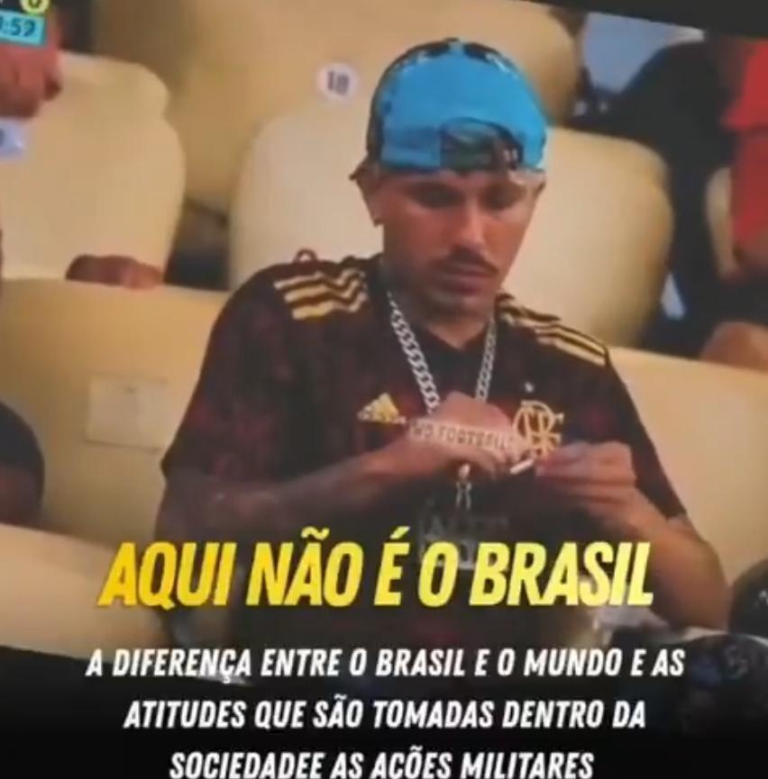 Uma fotografia exibe um homem de pele parda e cabelos escuros, com bigode e cavanhaque curtos, sentado em uma arquibancada com assentos beges. Ele veste um boné azul virado para trás, uma camisa esportiva em tons de vermelho escuro e preto com listras amarelas nos ombros e o escudo do time Flamengo, além de uma corrente prateada grossa no pescoço. O homem, que possui tatuagens visíveis nos braços e nas costas das mãos, olha para baixo enquanto manipula um cigarro entre os dedos. Sobreposta à parte inferior da imagem, há a frase 'AQUI NÃO É O BRASIL' em letras grandes e amarelas, seguida pelo texto em letras brancas menores: 'A DIFERENÇA ENTRE O BRASIL E O MUNDO E AS ATITUDES QUE SÃO TOMADAS DENTRO DA SOCIEDADEE AS AÇÕES MILITARES'.