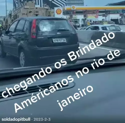 Uma fotografia capturada do interior de um veículo mostra uma cena de trânsito em uma via urbana. Em primeiro plano, nota-se o painel do carro do qual a foto foi tirada e, logo à frente, um automóvel escuro modelo hatch. À esquerda, parcialmente à frente do carro escuro, encontra-se um grande veículo blindado de cor bege com a inscrição 'PF-2' em preto na parte traseira. No extremo esquerdo da imagem, é possível ver parte de uma pessoa vestindo um colete refletivo amarelo, mas sem características físicas detalhadas discerníveis devido ao distanciamento e corte. Ao fundo, à direita, há um posto de combustíveis com o logotipo amarelo e vermelho da marca Shell e, à esquerda, um edifício com fachada de linhas diagonais pretas e brancas. Sobreposta à imagem, ocupando quase toda a metade inferior, uma grande frase em letras brancas inclinadas diz: 'chegando os Brindado Américanos no rio de janeiro'. No canto inferior esquerdo, lê-se em letras menores o texto 'soldadopitbull · 2023-2-3'.