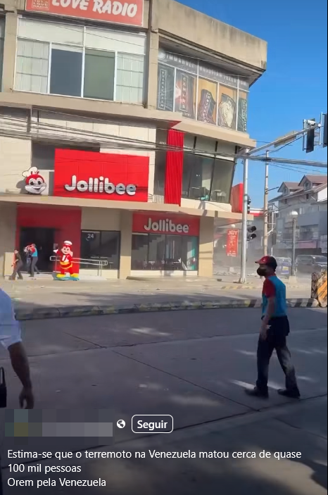 Uma captura de tela de um vídeo exibe uma cena de rua em frente a um edifício comercial de vários andares, onde se destaca um restaurante com grandes letreiros vermelhos com a inscrição 'Jollibee' e o desenho de seu mascote, uma abelha com chapéu de cozinheiro. Em primeiro plano, à direita, um indivíduo de pele parda — usando boné vermelho, máscara facial escura, camisa de mangas curtas azul e vermelha, calça escura e sapatos pretos —, caminha pela via. No canto esquerdo, é possível ver parcialmente o braço e parte do tronco de outra pessoa de pele parda vestindo uma blusa branca. Ao fundo, perto da entrada do estabelecimento onde há uma estátua do mascote, outras pessoas com trajes escuros aparecem desfocadas correndo ou caminhando rapidamente, enquanto uma densa nuvem de poeira cinza se ergue na rua do lado direito. No topo do prédio, há uma placa vermelha e branca com os dizeres 'LOVE RADIO'. Na parte inferior da imagem, sobreposta à cena, lê-se o texto em branco: 'Estima-se que o terremoto na Venezuela matou cerca de quase 100 mil pessoas Orem pela Venezuela', posicionado abaixo de um botão com a palavra 'Seguir' e um nome de usuário borrado digitalmente. 