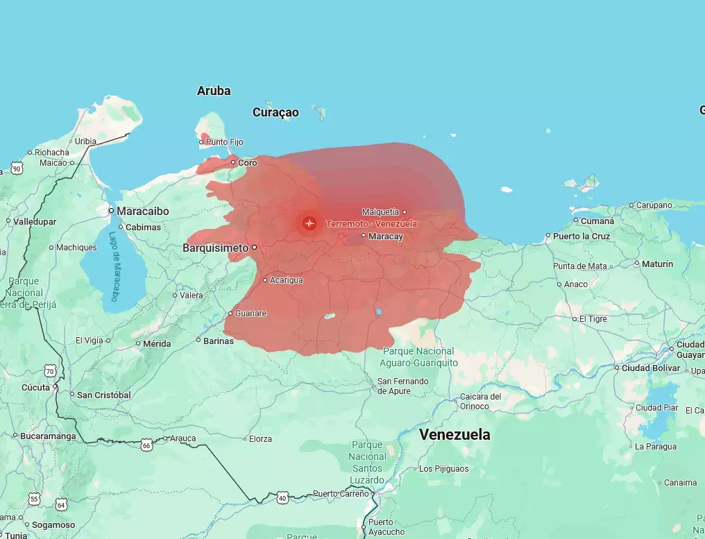 Uma imagem exibe uma reprodução de um mapa digital com foco na região norte da América do Sul, destacando o território da Venezuela. Sobre a porção centro-norte do país, estendendo-se do litoral em direção ao interior e cobrindo áreas próximas a cidades como Barquisimeto e Coro, há uma grande mancha vermelha semitransparente. No centro dessa área destacada, encontra-se um ícone circular vermelho com o desenho de uma onda sísmica branca, acompanhado pelo texto 'Terremoto • Venezuela'. A representação cartográfica apresenta o relevo terrestre em tons de verde e o oceano em azul-claro, identificando também ilhas caribenhas ao norte, como 'Aruba' e 'Curaçao'.