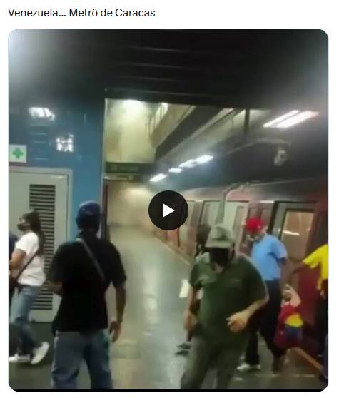 Uma captura de tela de uma postagem exibe o texto 'Venezuela... Metrô de Caracas' acima da miniatura de um vídeo que mostra a plataforma de uma estação de metrô. Ao fundo, uma densa nuvem de fumaça ou poeira clara preenche o espaço sob as luzes do teto, enquanto um trem com lataria vermelha e portas brancas encontra-se parado à direita. Na plataforma, circulam diversas pessoas, algumas utilizando máscaras faciais escuras. À esquerda, uma pessoa de cabelos escuros veste camiseta branca e calça jeans azul. Ao centro, de costas para a câmera, um indivíduo usa boné azul, camisa preta e calça jeans azul. Mais à direita, uma pessoa de pele clara veste boné verde, camisa de mangas curtas verde e calça escura, enquanto logo atrás, próximo ao trem, um homem de cabelos brancos veste boné vermelho, camisa azul-clara e calça escura. No centro da imagem, sobreposto à cena, há um ícone circular escuro com um símbolo de reprodução de vídeo em branco. 