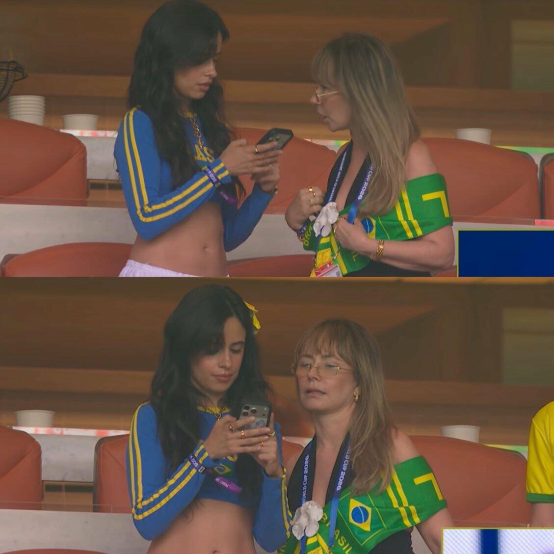 Uma imagem dividida em dois painéis horizontais exibe duas cenas sequenciais. Em ambos os quadros, duas mulheres estão posicionadas lado a lado em uma arquibancada com assentos avermelhados. A mulher à esquerda possui cabelos longos e escuros, veste uma blusa azul curta de mangas compridas com listras amarelas nos braços e detalhes em amarelo no peito, e segura um telefone celular. A mulher à direita tem pele clara, cabelos loiros com franja, usa óculos de grau e veste uma blusa preta coberta por um tecido verde e amarelo com formato e cores semelhantes aos da bandeira do Brasil, além de portar um cordão escuro de credencial no pescoço. No painel superior, a mulher de cabelo escuro segura o celular um pouco mais alto e olha para a tela, enquanto a mulher loira olha em direção a ela ou ao aparelho, segurando o tecido no peito; no painel inferior, a mulher de cabelo escuro abaixa levemente as mãos, continuando a olhar para o celular, enquanto a mulher loira direciona o olhar para a frente. 