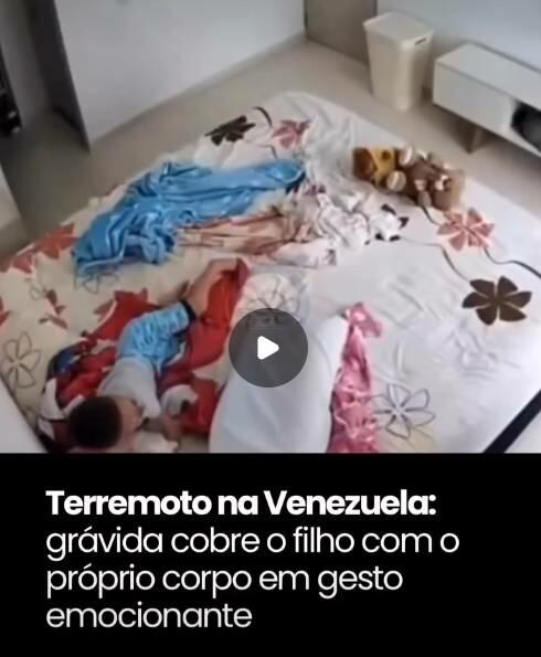 Uma captura de tela de um vídeo exibe uma cena de interior de uma residência, vista de um ângulo superior, mostrando uma grande cama ao centro. O lençol é claro com estampas de flores e folhas em tons escuros. Sobre a cama, do lado esquerdo, uma criança vestindo pijama azul repousa deitada, parcialmente coberta por uma manta vermelha com estampas azuis. Imediatamente ao seu lado, uma figura volumosa encontra-se inteiramente coberta por um tecido branco, posicionada de forma encurvada sobre a criança. Espalhados pela cama, há também um cobertor azul desarrumado, outras peças de tecido e um bicho de pelúcia marrom. No canto superior direito do enquadramento, observa-se o piso claro, uma lixeira branca e parte de um móvel branco com pés curtos de madeira. No centro da composição, sobreposto à imagem do quarto, há um ícone circular escuro com um símbolo de reprodução de vídeo em formato de triângulo branco. Na parte inferior, sobre uma ampla faixa preta sólida, há o texto em letras brancas: 'Terremoto na Venezuela:' em destaque, seguido logo abaixo por 'grávida cobre o filho com o próprio corpo em gesto emocionante'.
