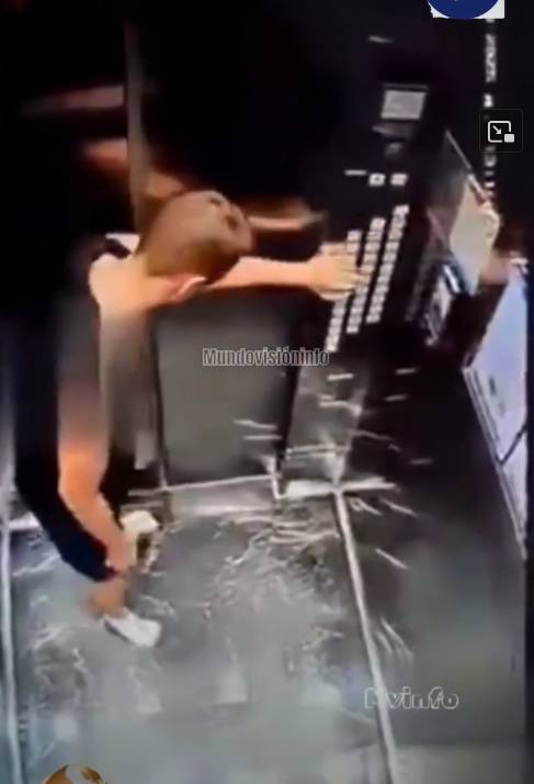 Uma captura de tela vertical, com aspecto de imagem de câmera de segurança, exibe o interior de um elevador visto de um ângulo superior. No centro da cena, um homem de pele clara e cabelos curtos, sem camisa e vestindo bermuda escura e tênis brancos, aparece de perfil. Ele está com o braço direito estendido para a frente, tocando a parte superior de um grande painel de botões escuro localizado na parede direita. Sua mão esquerda segura um pequeno objeto claro na altura da cintura. Uma faixa horizontal com efeito de desfoque digital atravessa a região do seu peito e as costas. O piso do elevador é escuro, com marcas e reflexos claros que lembram rachaduras ou um padrão marmorizado, enquanto a parede ao fundo apresenta um tom quente amadeirado ou alaranjado. Sobreposta ao centro da imagem, há uma marca d'água translúcida com o texto 'MundoVisiónInfo', e no canto inferior direito, outra marca d'água diz 'Mvinfo'. No canto superior direito, notam-se caracteres brancos desfocados típicos de registros de data e hora de monitoramento, além de um ícone de tela cheia. 