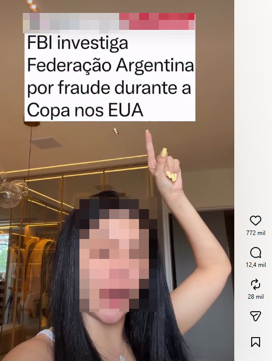 Uma captura de tela vertical de um vídeo em rede social exibe em primeiro plano uma mulher de longos cabelos escuros e blusa branca, cujo rosto está totalmente oculto por um efeito quadriculado (pixelizado). Ela mantém o braço direito erguido, apontando o dedo indicador para o alto, exibindo unhas longas pintadas de amarelo claro. Acima dela, na região para onde aponta, há um grande bloco retangular branco contendo o texto em letras pretas: ‘FBI investiga Federação Argentina por fraude durante a Copa nos EUA’. A borda superior desse bloco possui uma estreita faixa horizontal borrada digitalmente que mescla tons de cinza e vermelho. O fundo da cena revela um ambiente interno com iluminação quente, teto com luzes embutidas e um móvel amplo que se assemelha a um closet com portas de vidro e prateleiras iluminadas por fitas de LED. No lado direito da tela, alinhados verticalmente, estão os ícones de interação típicos da plataforma, acompanhados de seus respectivos números: um coração com ‘772 mil’, um balão de comentário com ‘12,4 mil’, um símbolo de repetição com ‘28 mil’, seguidos por ícones de compartilhar e salvar. 