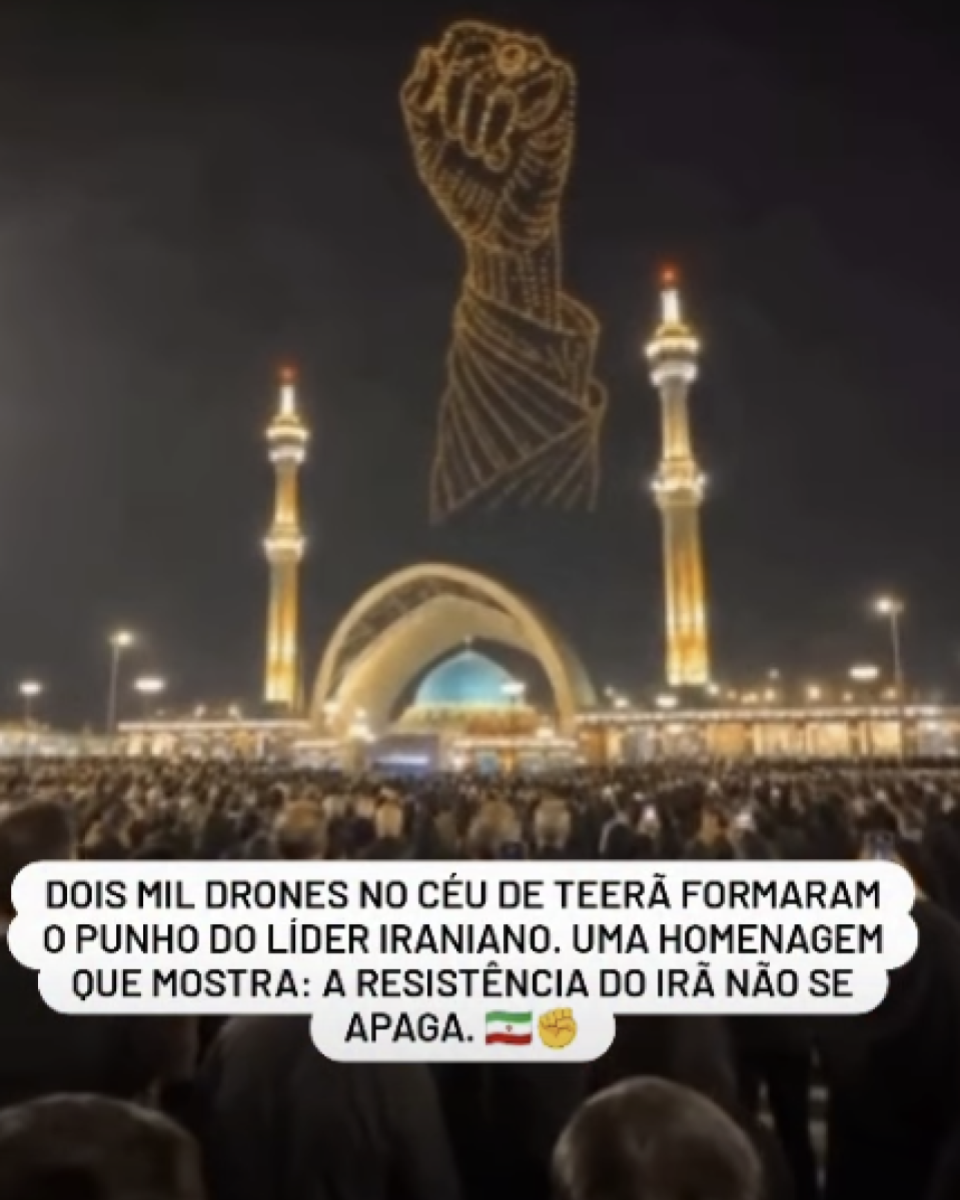 A imagem mostra uma cena noturna em frente a um grande templo iluminado, com duas torres altas e douradas posicionadas uma de cada lado de uma estrutura em forma de arco. Uma grande multidão ocupa toda a área em primeiro plano, vista de costas, voltada para o templo. Acima da construção, no céu escuro, centenas de pontos luminosos formam o desenho de um punho cerrado, contornado por luzes douradas. Na parte inferior da imagem há uma caixa de texto branca com cantos arredondados. Nela está escrito: 'DOIS MIL DRONES NO CÉU DE TEERÃ FORMARAM O PUNHO DO LÍDER IRANIANO. UMA HOMENAGEM QUE MOSTRA: A RESISTÊNCIA DO IRÃ NÃO SE APAGA.', seguida de um emoji da bandeira do Irã e um emoji de punho cerrado.