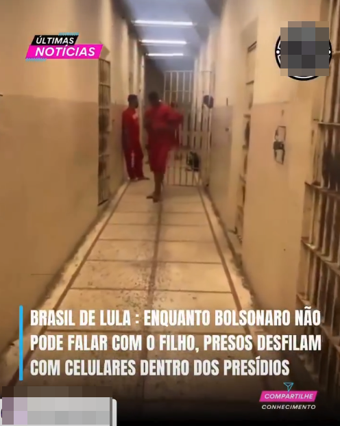 Uma captura de tela vertical de um vídeo exibe a visão de um corredor comprido e estreito, característico de um presídio, com piso claro demarcado por linhas e portas de celas com grades metálicas em ambos os lados, sob iluminação artificial no teto. Ao fundo da passagem, duas pessoas vestindo uniformes compostos por calça e camisa vermelhas soltas movimentam-se pelo corredor. Ocupando a parte inferior da tela, em letras maiúsculas brancas alinhadas à esquerda junto a uma barra vertical azul, lê-se a seguinte frase: ‘BRASIL DE LULA : ENQUANTO BOLSONARO NÃO PODE FALAR COM O FILHO, PRESOS DESFILAM COM CELULARES DENTRO DOS PRESÍDIOS’. Logo abaixo, no canto inferior direito, um pequeno ícone triangular azul é acompanhado pelas palavras ‘COMPARTILHE’ dentro de uma pequena tarja rosa, e ‘CONHECIMENTO’ em letras menores brancas logo abaixo. Por fim, no canto inferior esquerdo, há uma área retangular horizontal que foi borrada digitalmente.