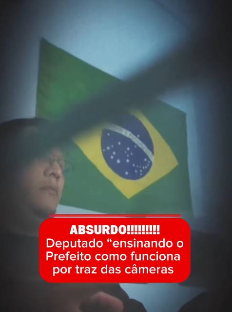 Uma captura de tela de um vídeo vertical em formato de rede social, com um feixe de luz cruzando a cena escura. No lado esquerdo, vê-se o perfil de um homem de óculos, e atrás dele está pendurada ou projetada uma bandeira do Brasil. Sobreposto na parte inferior, há um quadro vermelho retangular com texto em branco em português: ‘ABSURDO!!!!!!!!!!!’ na primeira linha, seguido por ‘Deputado 'ensinando o Prefeito como funciona por traz das câmeras’ nas linhas seguintes. 
