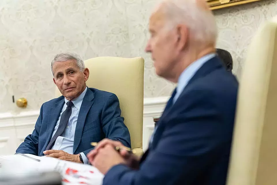 Uma imagem de média distância captura Anthony Fauci e Joe Biden sentados em poltronas bege de encosto alto em um escritório que se assemelha ao Salão Oval. Fauci, um homem de pele clara e cabelo curto grisalho, está em foco e olha para a direita, em direção a Biden. Ele veste um terno azul-marinho com um padrão xadrez sutil, uma camisa social azul-clara e uma gravata escura texturizada. Biden, posicionado em primeiro plano à direita e desfocado, é um homem de pele clara com pouco cabelo grisalho, usando um paletó azul-marinho. Suas mãos estão visíveis no canto inferior, segurando uma caneta sobre alguns papéis desfocados. O fundo mostra uma parede com papel de parede de padrão claro e rodapés brancos.