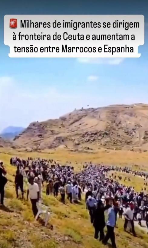 A fotografia exibe uma ampla multidão caminhando por um terreno montanhoso e irregular, coberto por vegetação rasteira de tom amarelo-acastanhado. As pessoas, capturadas a certa distância, apresentam tons de pele variados e majoritariamente cabelos escuros, vestindo trajes diversos que incluem camisas brancas, azuis e pretas, calças escuras e chapéus. Uma das pessoas no canto inferior esquerdo carrega uma cadeira de plástico branca. O céu ao fundo é azul e claro. Sobreposta na parte superior da imagem, há uma caixa de fundo branco contendo um emoji de sirene vermelha e o seguinte texto em letras escuras: 'Milhares de imigrantes se dirigem à fronteira de Ceuta e aumentam a tensão entre Marrocos e Espanha'. 