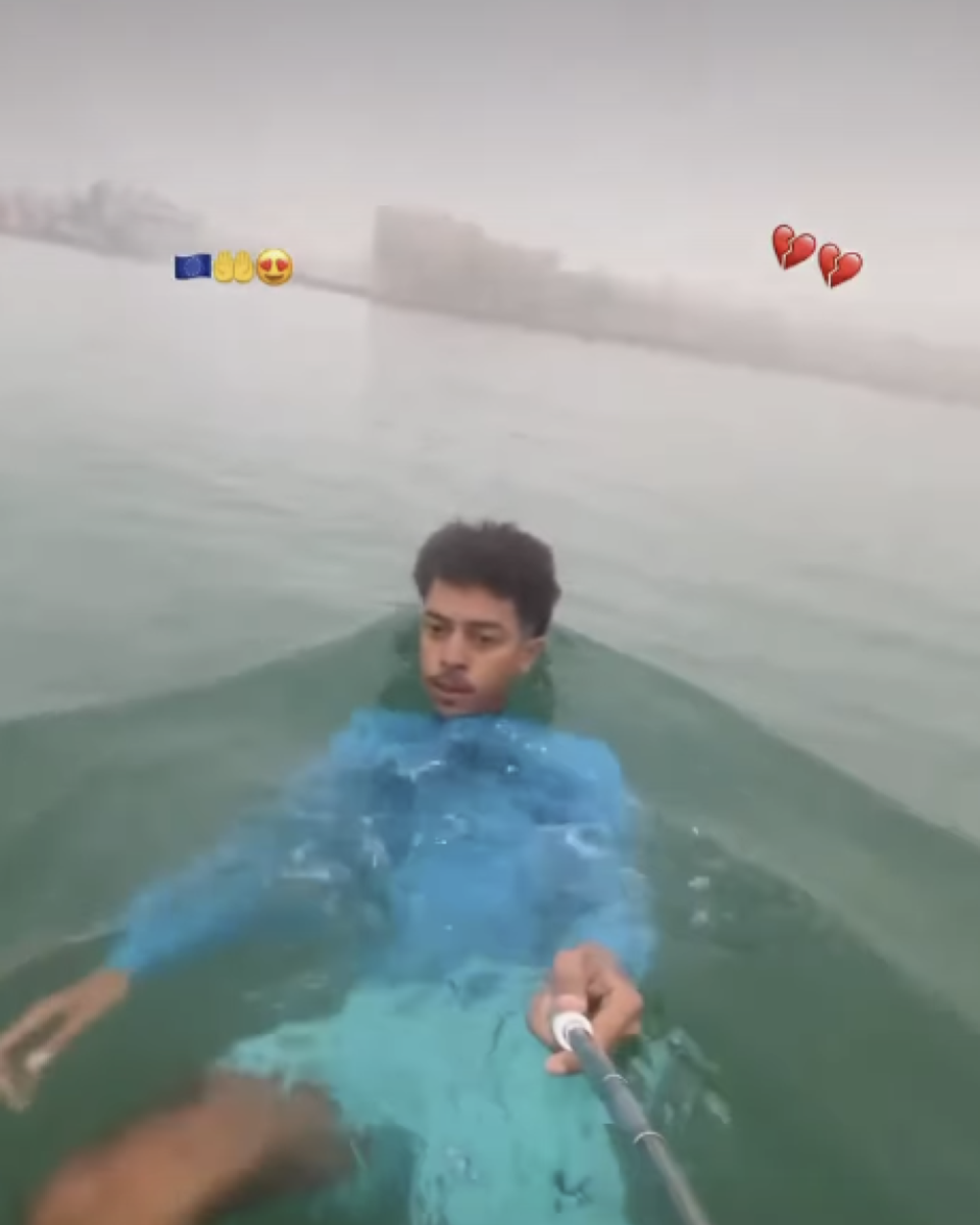 A imagem mostra uma pessoa jovem nadando, com a água chegando aproximadamente à altura do peito. Ela tem cabelos escuros e curtos e veste uma camisa azul-clara e uma peça de roupa verde-clara. A pessoa está segurando um bastão de selfie com a mão direita, enquanto mantém o braço esquerdo estendido para o lado. Ao fundo, há uma parede clara e uma estrutura retangular pouco definida. Na parte superior da imagem aparecem um emoji da bandeira da União Europeia, dois emojis de mãos amarelas e um emoji de rosto sorridente com olhos em forma de coração, enquanto no lado direito há dois emojis de coração partido.