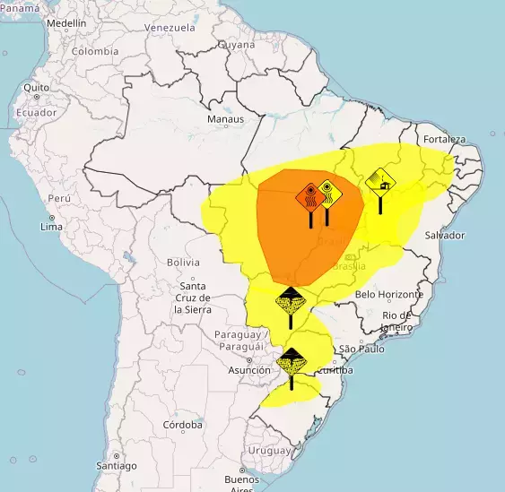 Mapa da América do Sul com foco no território brasileiro, apresentando manchas coloridas sobrepostas que indicam áreas de alerta. Uma extensa zona amarela abrange porções das regiões Nordeste, Centro-Oeste, Sudeste e Sul do Brasil, contendo em seu interior uma área menor de cor laranja concentrada predominantemente no Centro-Oeste. Sobre essas manchas, há cinco ícones em formato de losango simulando placas de sinalização fixadas por hastes: na região central, vê-se uma placa laranja e uma amarela com símbolos de radiação e calor (sol e linhas onduladas ascendentes), além de uma placa amarela com o desenho do que aparenta ser uma fábrica ou medidor de umidade; mais ao sul, ao longo da faixa amarela, encontram-se duas placas amarelas ilustrando nuvens carregadas com precipitação. Nomes de cidades, como 'Brasília' e 'São Paulo', estão grafados no mapa base.