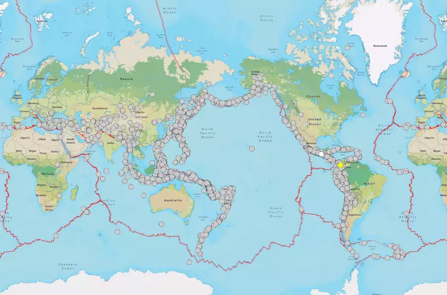 A imagem exibe um mapa-múndi planificado com continentes em tons de verde e marrom e oceanos em azul-claro. Linhas vermelhas irregulares atravessam a superfície global, delineando diversas áreas geográficas. Centenas de pequenos círculos cinzas estão agrupados majoritariamente ao longo e nas proximidades dessas linhas vermelhas, formando padrões densos especialmente nas bordas do Oceano Pacífico, no sul da Ásia e na região do Mar Mediterrâneo. Nomes de países, continentes e oceanos aparecem escritos em letras pequenas sobre o mapa.