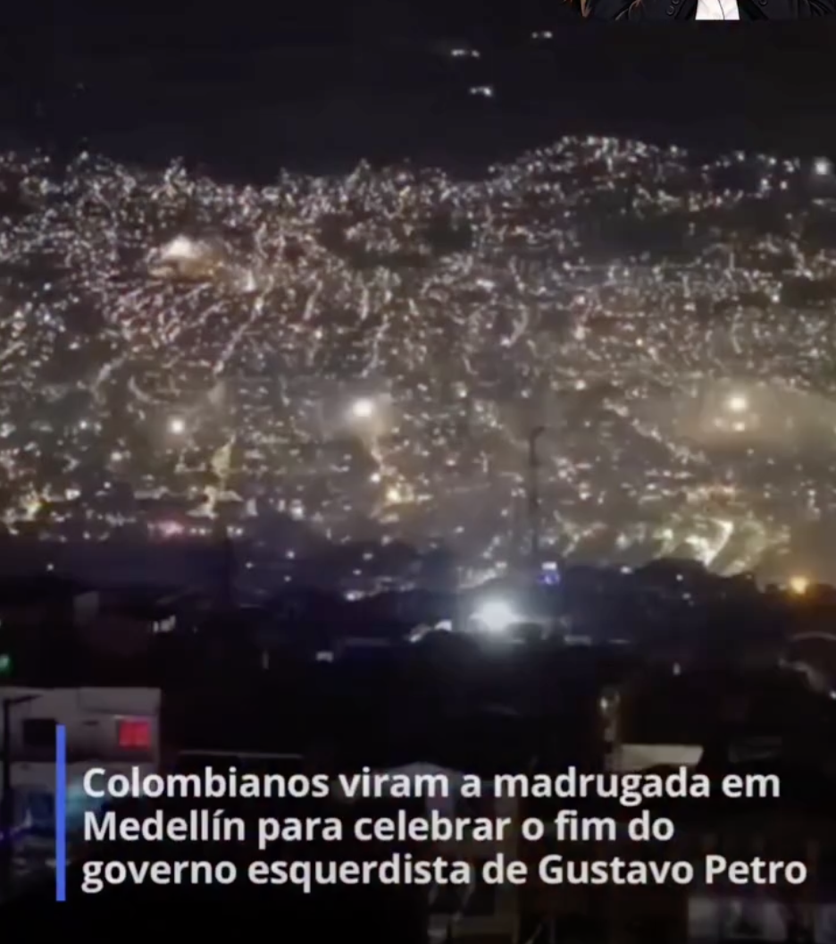 Imagem mostra trecho de um vídeo gravado durante à noite de um local alto, que mostra uma vista panorâmica da cidade de Medellín, na Colômbia. A paisagem é montanhosa e iluminada. Fogos de artifício coloridos explodem no céu noturno enquanto estrondos contínuos são ouvidos. Na parte inferior, uma caixa de texto azul e branca traz a frase: 