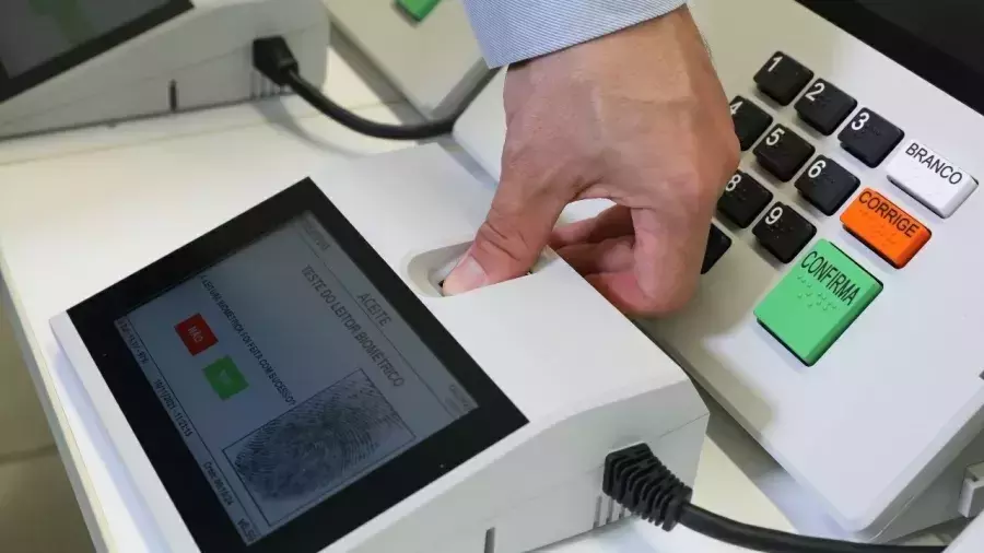 Foto exibe o detalhe da mão direita de uma pessoa de pele clara — vestindo uma camisa de mangas compridas azul-clara com listras finas —, posicionando o polegar sobre o sensor de um leitor biométrico de mesa. O dispositivo branco, conectado por um cabo preto, apresenta uma tela inclinada que mostra a imagem gráfica de uma impressão digital em tons de cinza e os textos 'ACEITE' e 'TESTE DO LEITOR BIOMÉTRICO', além de marcações em vermelho e verde. À direita do leitor, é visível uma porção do painel de uma urna eletrônica contendo teclas numéricas pretas e três teclas de comando sobrepostas verticalmente: uma branca com a inscrição 'BRANCO', uma laranja com 'CORRIGE' e uma verde com 'CONFIRMA'.