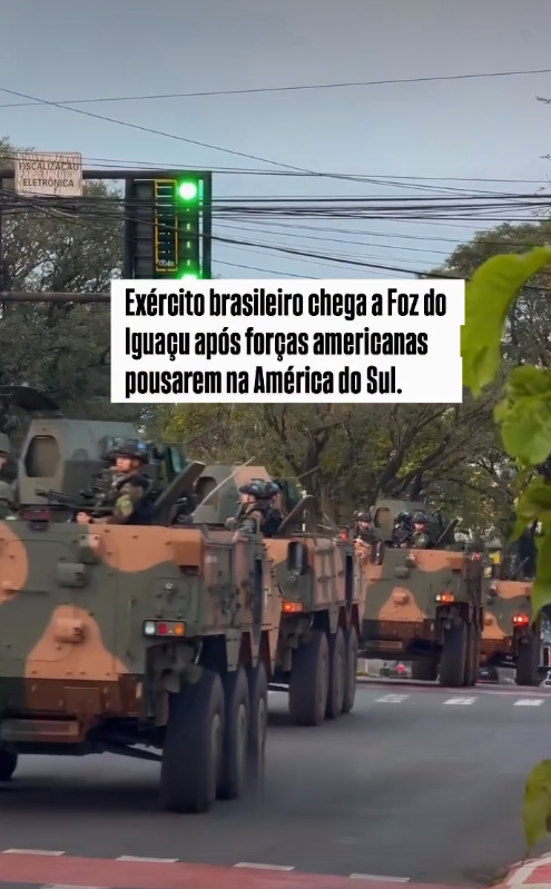 A imagem exibe um comboio de veículos militares blindados de seis rodas, com pintura camuflada em tons de verde, marrom e preto, transitando em fila por uma via urbana asfaltada. Nas escotilhas superiores dos veículos, é possível observar soldados — homens de pele clara, vestindo uniformes camuflados e capacetes escuros. O cenário urbano inclui árvores ao fundo, fiação elétrica aérea, um semáforo com a luz verde acesa e uma placa com a inscrição 'FISCALIZAÇÃO ELETRÔNICA'. Sobreposta à parte central superior da imagem, há uma caixa de fundo branco contendo o seguinte texto em letras pretas: 'Exército brasileiro chega a Foz do Iguaçu após forças americanas pousarem na América do Sul.'. No canto inferior direito, ramificações de folhas verdes aparecem desfocadas em primeiro plano.