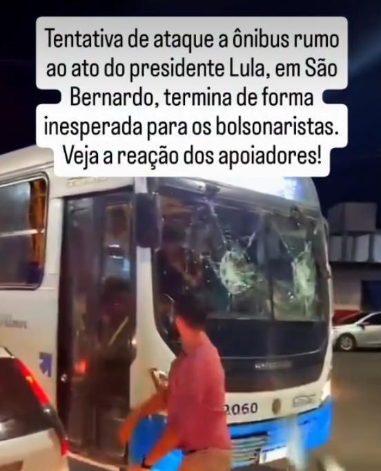 Esta imagem mostra um homem com uma camisa rosa avermelhada que está de pé na frente de um ônibus. O veículo é branco com para-choques azuis e vários vidros da parte frontal e lateral do ônibus estão visivelmente quebrados, com buracos e rachaduras. Na parte superior da imagem, um bloco de texto grande e branco é sobreposto à cena com a seguinte legenda: ‘Tentativa de ataque a ônibus rumo ao ato do presidente Lula, em São Bernardo, termina de forma inesperada para os bolsonaristas. Veja a reação dos apoiadores!’