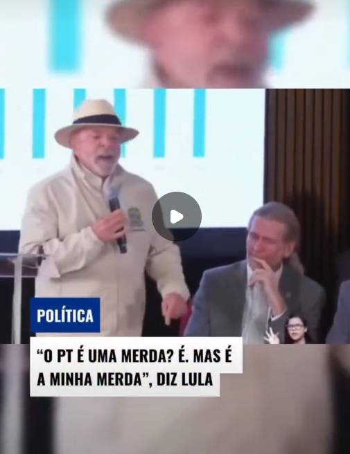 Captura de tela de um vídeo com um botão de reprodução escuro sobreposto ao centro. Em destaque à esquerda, está Luiz Inácio Lula da Silva — homem de pele clara e barba grisalha —, vestindo um chapéu claro com faixa escura e uma jaqueta bege, segurando um microfone com a mão direita. Sentado à direita, observa-se um homem de pele clara e cabelo grisalho comprido amarrado, usando terno cinza e gravata clara, com a mão direita repousada próxima ao rosto. No canto inferior direito, um pequeno quadro sobreposto exibe uma intérprete de libras — mulher de pele clara, cabelo escuro e óculos —, vestindo blusa preta. O fundo mostra uma tela branca com gráficos de barras verticais em azul-claro. Na parte inferior esquerda, há uma etiqueta azul com o texto 'POLÍTICA' em letras brancas, e logo abaixo, um letreiro branco com a seguinte inscrição em letras maiúsculas pretas: 'O PT É UMA MERDA? É. MAS É A MINHA MERDA', DIZ LULA. 