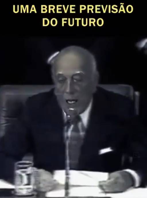 Imagem exibe, em tom predominantemente acinzentado, um homem idoso de pele clara, calvo no topo da cabeça e com cabelos claros nas laterais. Ele veste um terno escuro com um lenço claro no bolso do peito, camisa branca e gravata texturizada, e aparece com a boca entreaberta, posicionado diante de um microfone fino sobre uma mesa onde repousam papéis e um copo de água à sua esquerda. Na parte superior da imagem, contrastando com o fundo escuro, lê-se o texto em letras maiúsculas amarelas: 'UMA BREVE PREVISÃO DO FUTURO'.