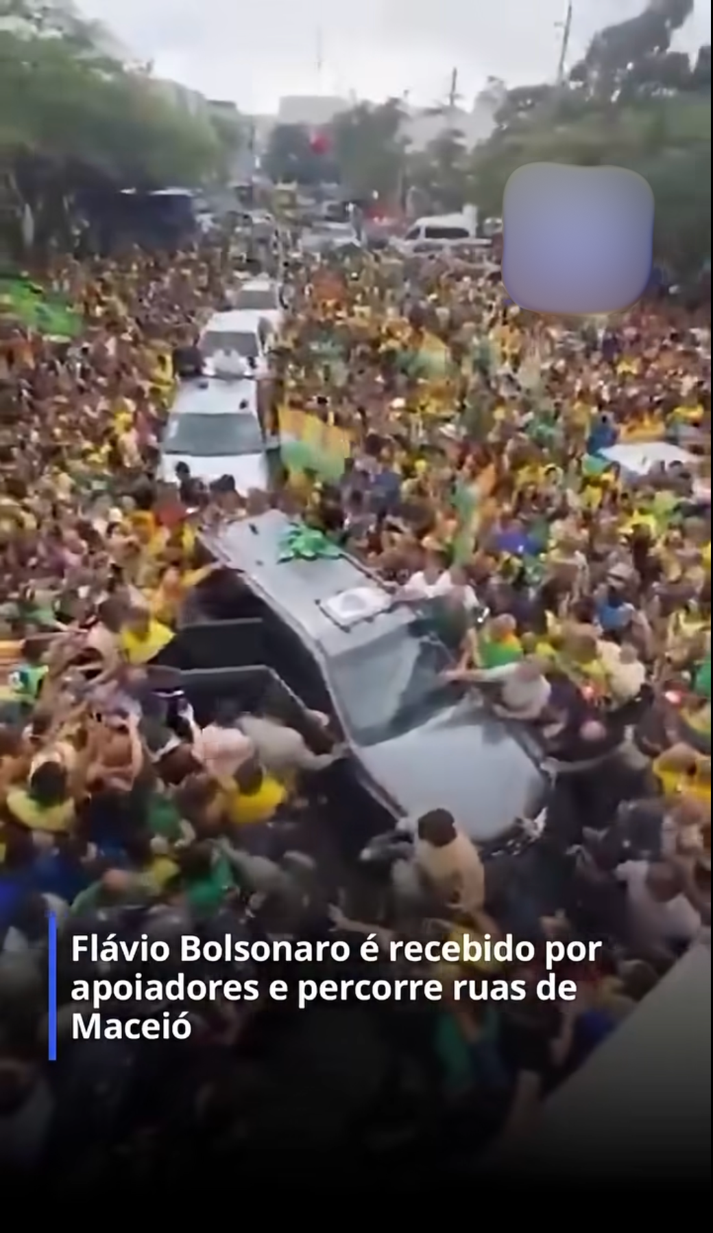 Uma vista aérea de um grande ato político em uma via pública movimentada. Uma grande multidão de apoiadores, em sua maioria vestindo roupas amaral e verde, cerca completamente um veículo SUV escuro que avança devagar no centro da rua. Pessoas cercam o carro tentando se aproximar, e ao fundo é possível ver outros veículos da carreata parados na avenida tomada pela multidão. Na parte inferior da imagem, há uma caixa de legenda com texto em letras brancas sobre fundo escuro: ‘Flávio Bolsonaro é recebido por apoiadores e percorre ruas de Maceió’.