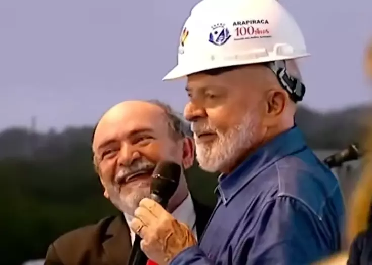 O presidente Luiz Inácio Lula da Silva — homem de pele clara, cabelos e barba brancos — aparece em primeiro plano vestindo uma camisa social azul e um capacete de proteção branco que exibe logotipos e a inscrição ‘ARAPIRACA 100 anos’. Ele segura um microfone preto próximo ao rosto com a mão direita. Logo atrás, à sua esquerda, está um segundo homem de pele clara, calvo com cabelos brancos nas laterais e barba branca curta, que sorri vestindo um paletó escuro sobre uma camisa branca. O fundo da imagem é desfocado, mostrando um céu claro e silhuetas de árvores em um ambiente externo.