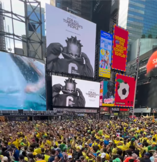 ALT text: Uma grande multidão ocupa uma praça cercada por enormes painéis de publicidade e prédios altos, na Times Square, em Nova York. A maioria das pessoas veste roupas amarelas e verdes. No centro superior, um grande painel branco exibe a imagem de uma criança negra com tranças, usando uma camiseta preta, acompanhada da mensagem For All Tomorrow’s People; no telão em vermelho ao lado há a imagem de uma bola de futebol e escrito em inglês ‘onde o mundo vem jogar’.