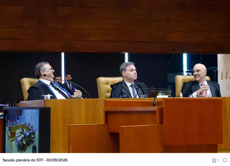 Três homens de pele clara, vestindo togas pretas sobre camisas brancas e gravatas, estão sentados lado a lado em cadeiras de couro claro atrás de uma ampla bancada de madeira. Flávio Dino, posicionado à esquerda possui cabelos grisalhos, usa óculos e sorri olhando para o lado. André Mendonça, ao centro, tem cabelos curtos escuros com tons grisalhos, olhar direcionado para a frente e mantém uma expressão séria. Alexandre de Moraes, à direita, é calvo e também sorri, olhando na direção do centro da bancada. Ao fundo, parcialmente desfocados, dois homens em trajes sociais aparecem observando a sessão. Em primeiro plano, no canto inferior esquerdo, a tela de um monitor exibe a gravação com a visão panorâmica do plenário. Na parte inferior da imagem, um texto sobre fundo branco informa: 'Sessão plenária do STF - 03/09/2026'.