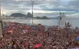 Imagem da Praia de Copacabana vista de frente para o mar, com uma multidão muito grande de pessoas ocupando a faixa de areia. Ao fundo, aparecem o mar, montanhas e prédios da orla, enquanto algumas bandeiras, guarda-sóis e estruturas se destacam no meio do público.